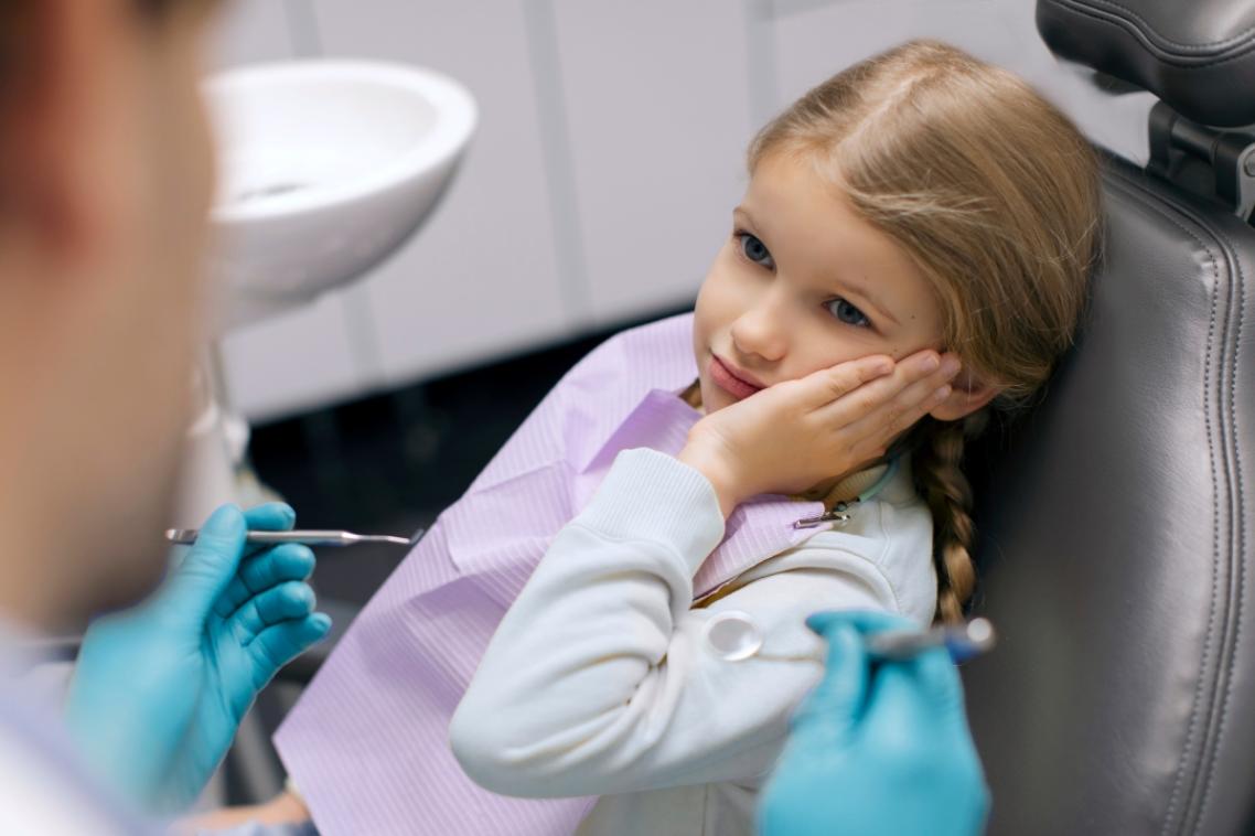 A young girl covers the side of her face while sitting in a dentist's chair.